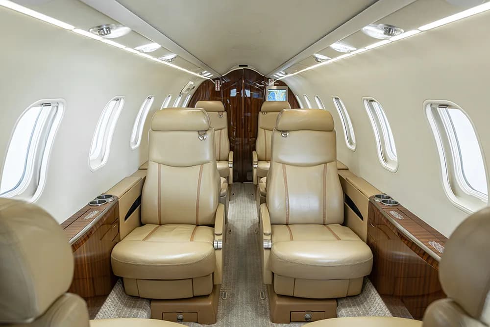 A view down the aisle of a private jet cabin with tan leather seats and wood accents.