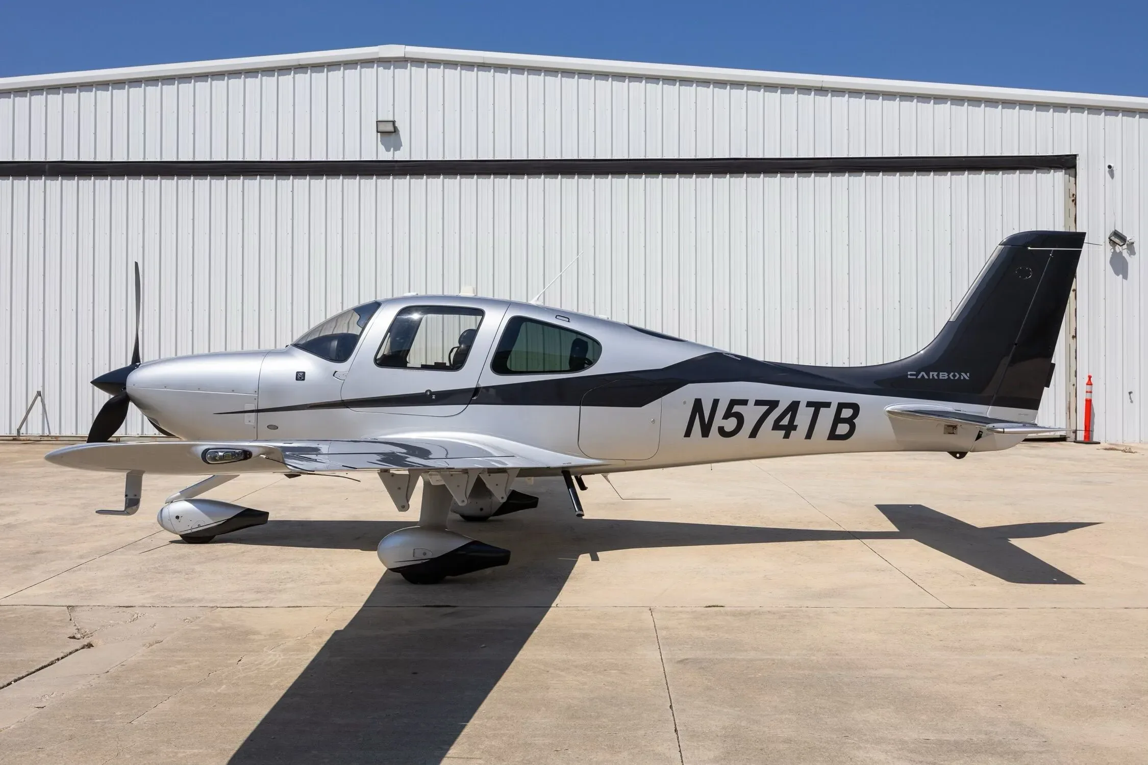 A clear side profile of the silver and black Cirrus SR22 aircraft, registration N574TB, against a white hangar.