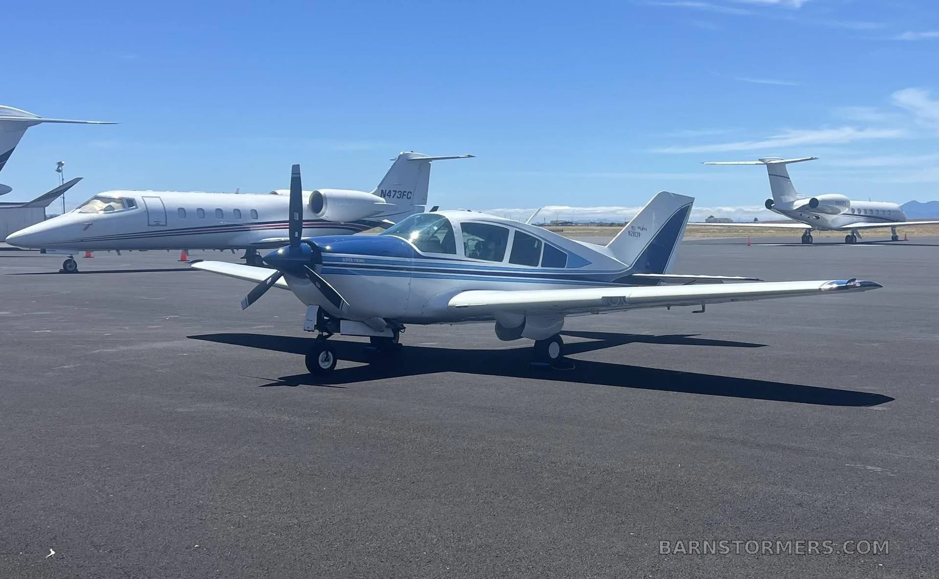 A white and blue Bellanca Viking aircraft parked on an airport tarmac with other private jets under a clear blue sky.