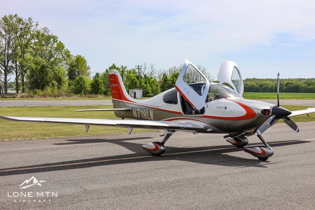 Three-quarter front view of a silver and orange Cirrus SR22 aircraft with both doors open, parked on an asphalt runway.