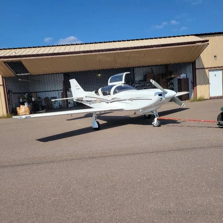 A white Vans RV-10 or similar kit plane with black and grey stripes, being towed out of a hangar on a sunny day.