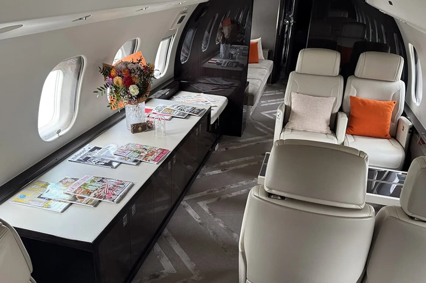 A wide view of a private jet's luxurious cabin, featuring a long credenza with magazines and a floral arrangement, and multiple seating areas.