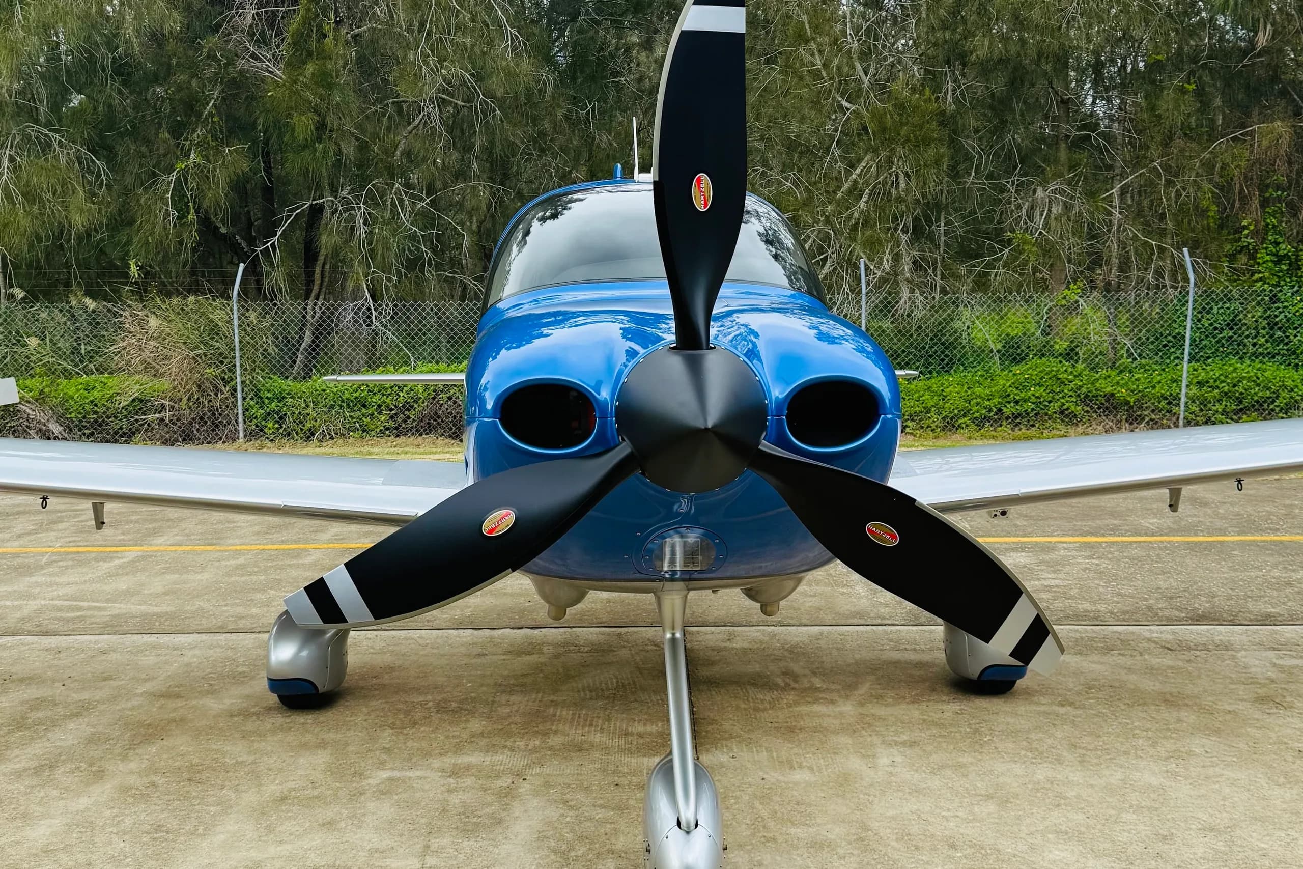 A front view of a blue and silver single-engine propeller aircraft with a black Hartzell propeller, parked on a concrete tarmac.