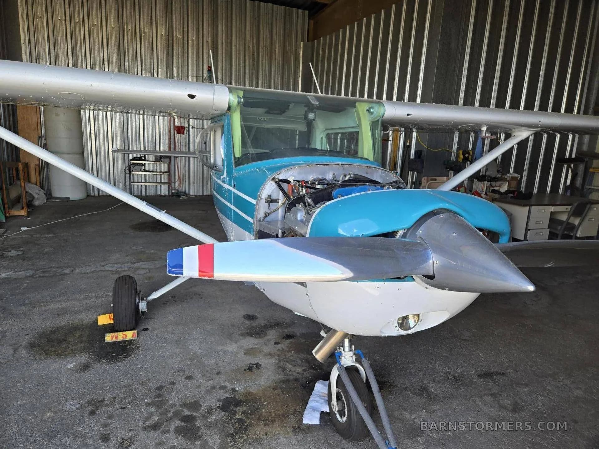 A front-quarter view of a light aircraft in a hangar, with the engine partially exposed and a teal and white paint scheme.