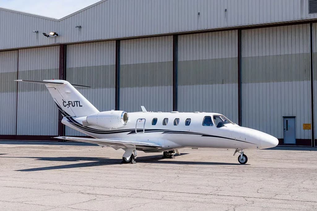 A white Cessna Citation business jet with black and grey stripes, registration C-FUTL, parked in front of a large hangar.
