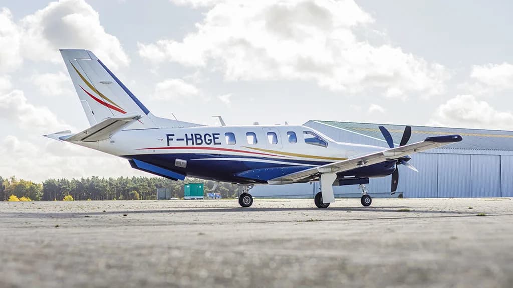 A white and blue TBM aircraft with registration F-HBGE parked on an airfield with a large hangar in the background.