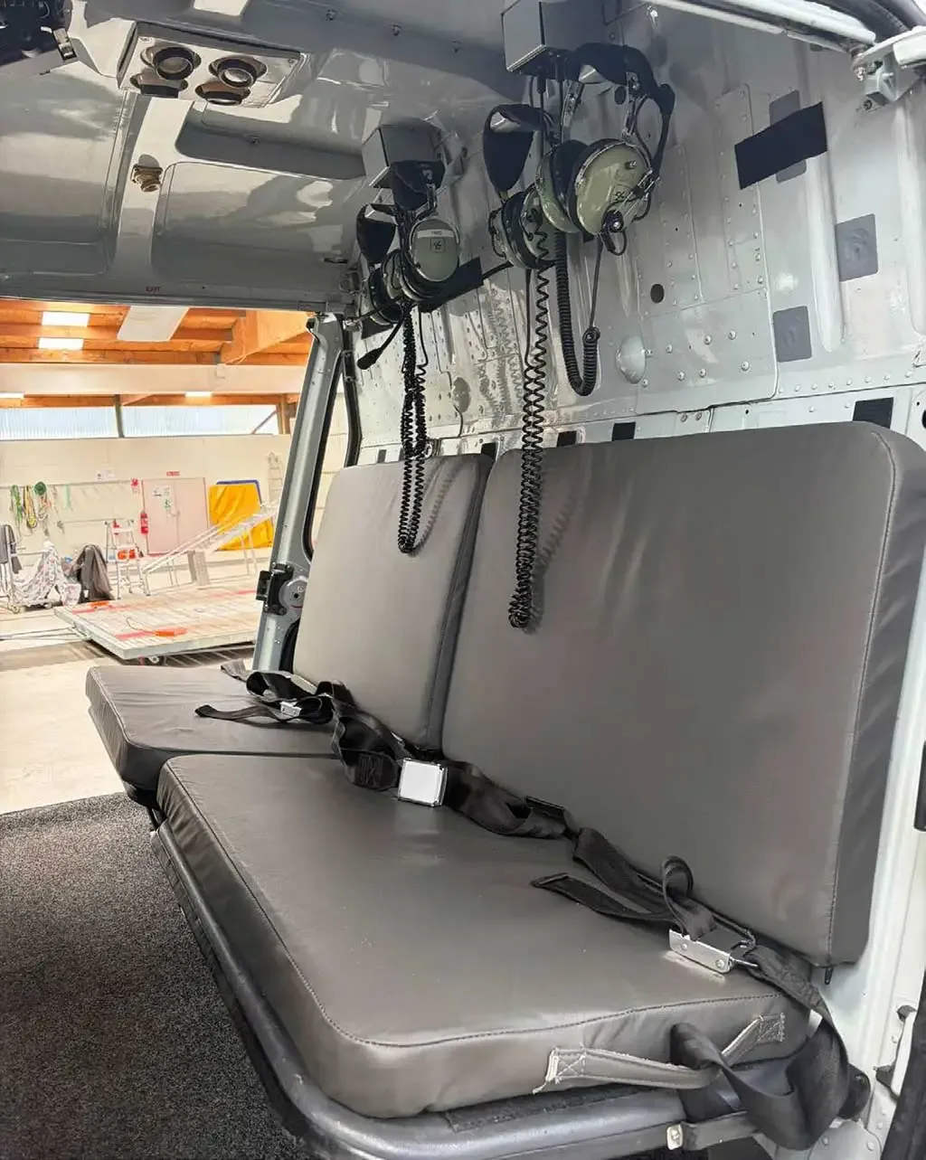 The grey rear passenger seats of a helicopter cabin with headsets hanging above.