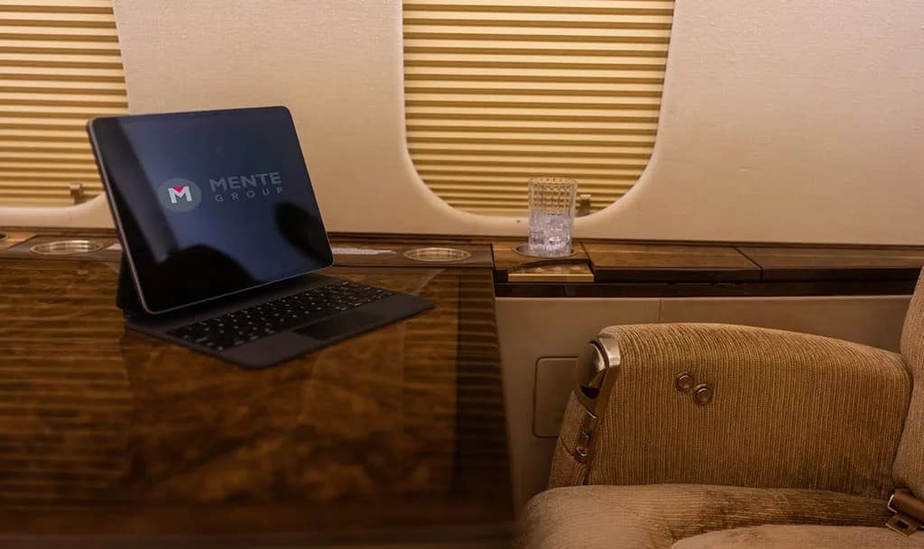 A close-up view of a polished dark wood table inside a private jet, with a tablet displaying the "Mente Group" logo and a glass of water.