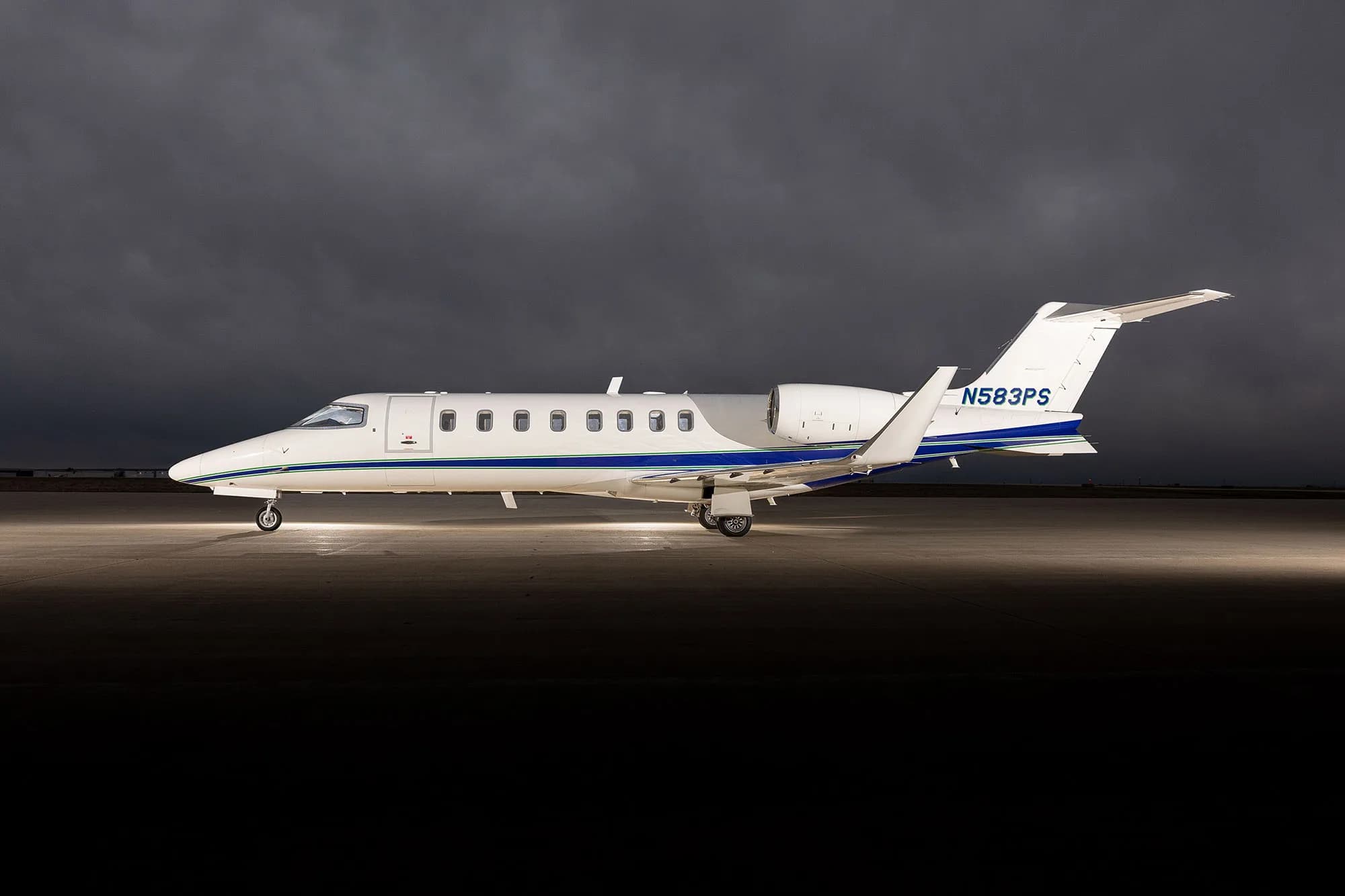 A white Learjet 75 with blue and green stripes, registration N583PS, parked on an tarmac under dramatic cloudy skies.