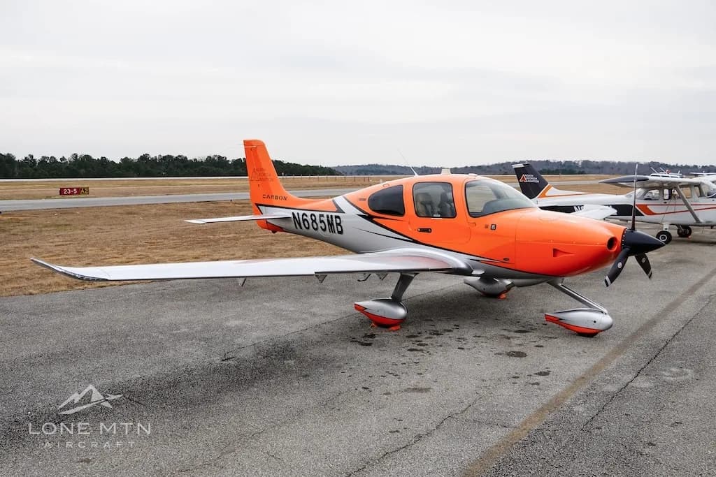The orange and silver Cirrus SR20 aircraft N685MB parked on an airfield with other planes in the distance.