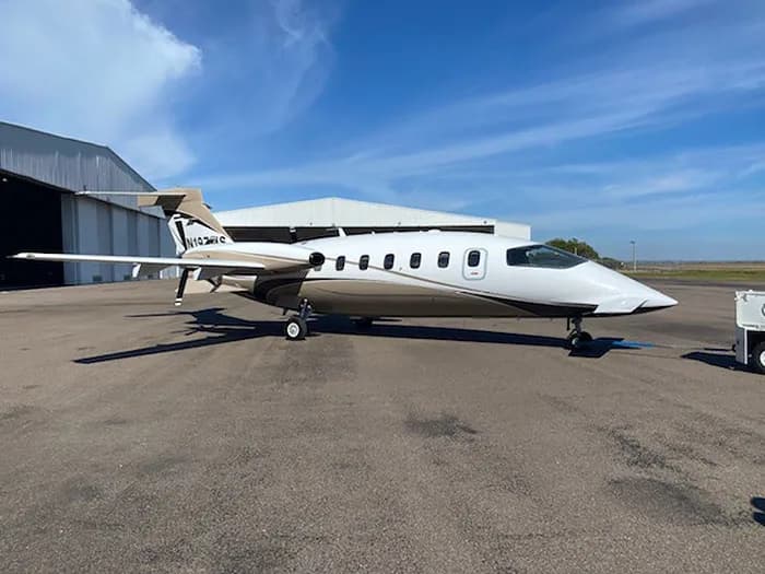 A white and tan Piaggio P.180 Avanti aircraft parked on the tarmac next to a hangar.