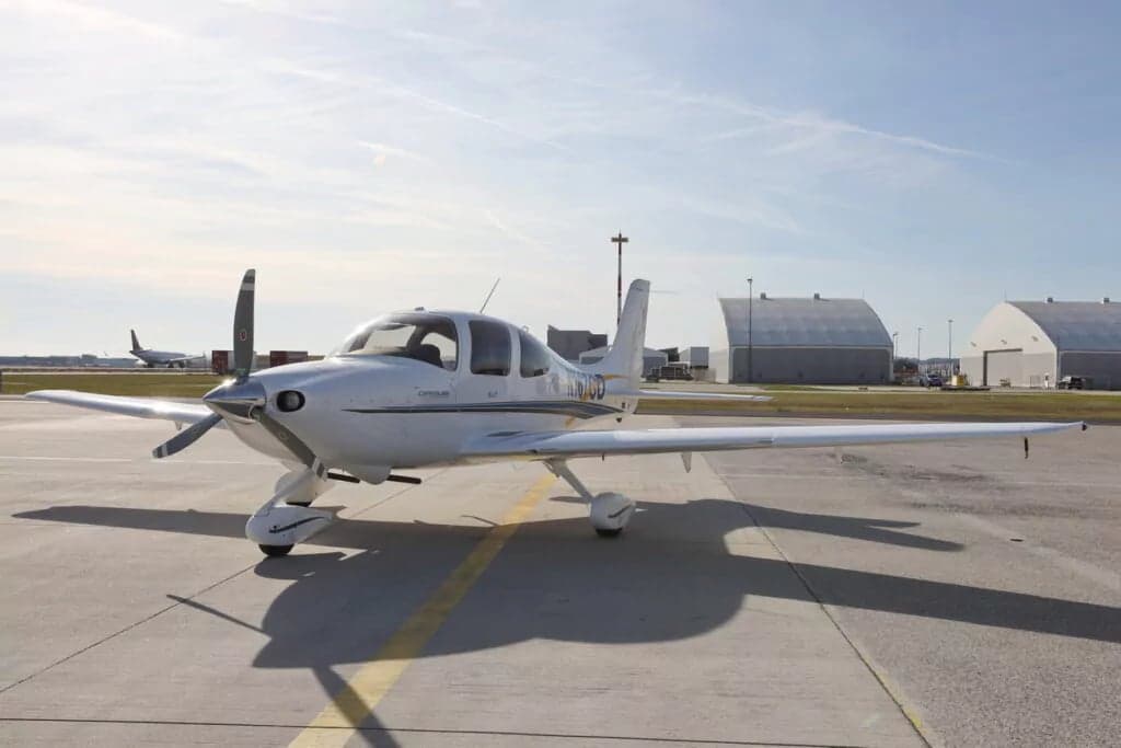 A Cirrus SR22 aircraft parked on the tarmac with hangars in the background.