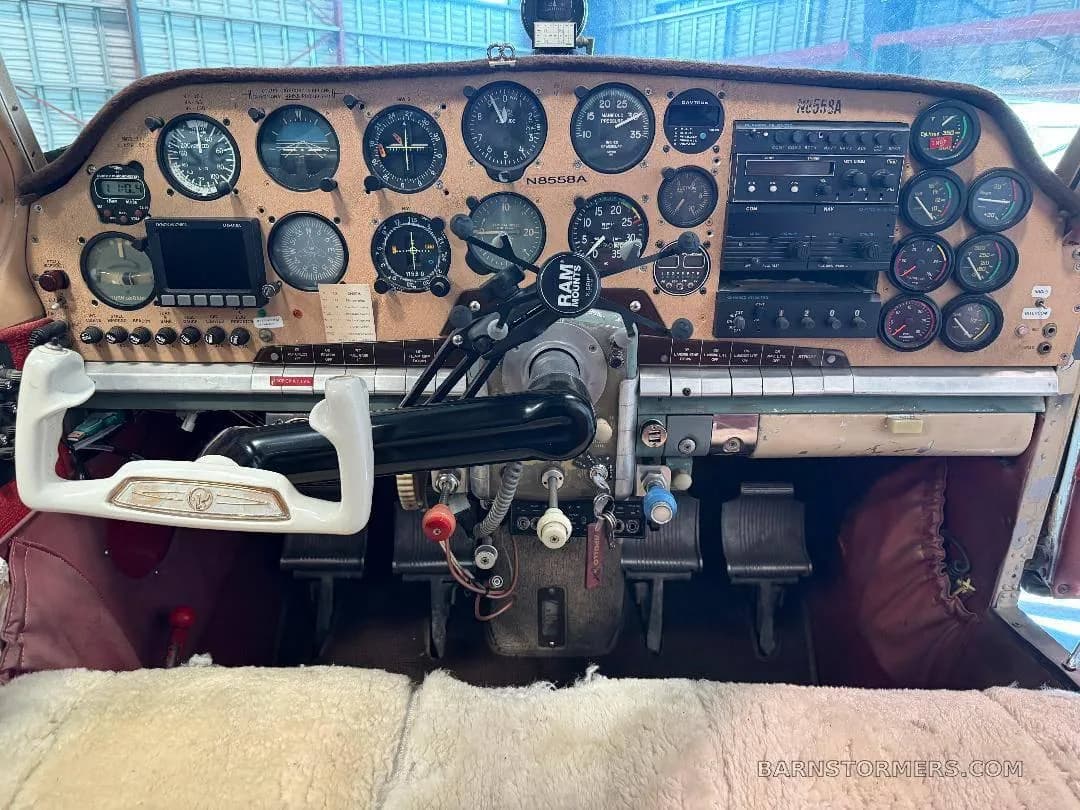 The cockpit of a vintage aircraft features a brown instrument panel with analog gauges, a white yoke, and red fabric interior.