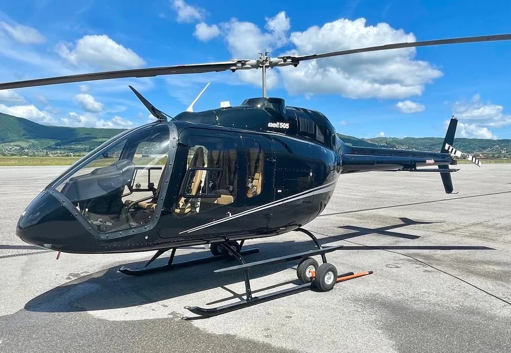 A black Bell 505 helicopter with tan interior on an airport tarmac under a blue sky.