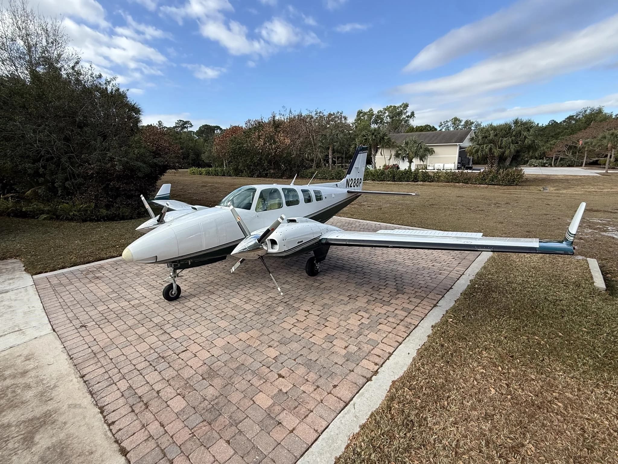 A white and green twin-engine Beechcraft Baron aircraft with tail number N288P parked on a brick driveway under a blue sky.
