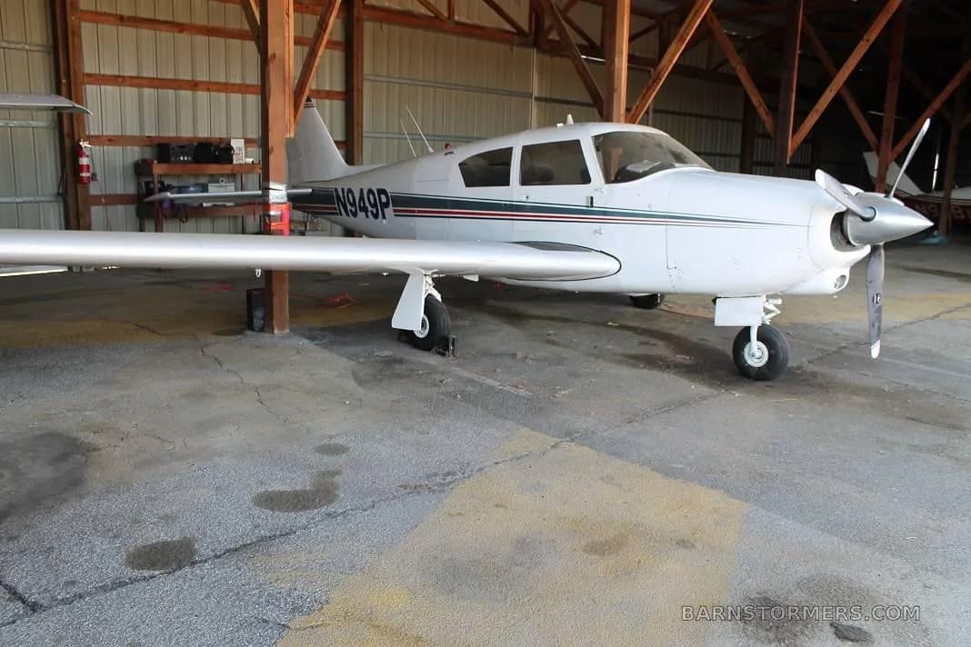 A white single-engine propeller aircraft with blue and red stripes, registration N949P, parked inside a wooden hangar.