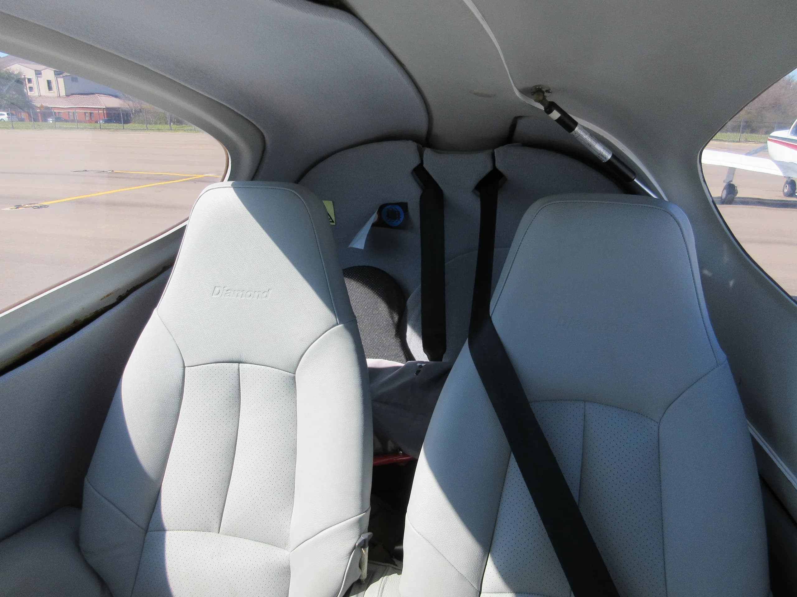 The light gray interior seats of a Diamond aircraft, viewed from the cockpit looking back, with an airport visible outside.