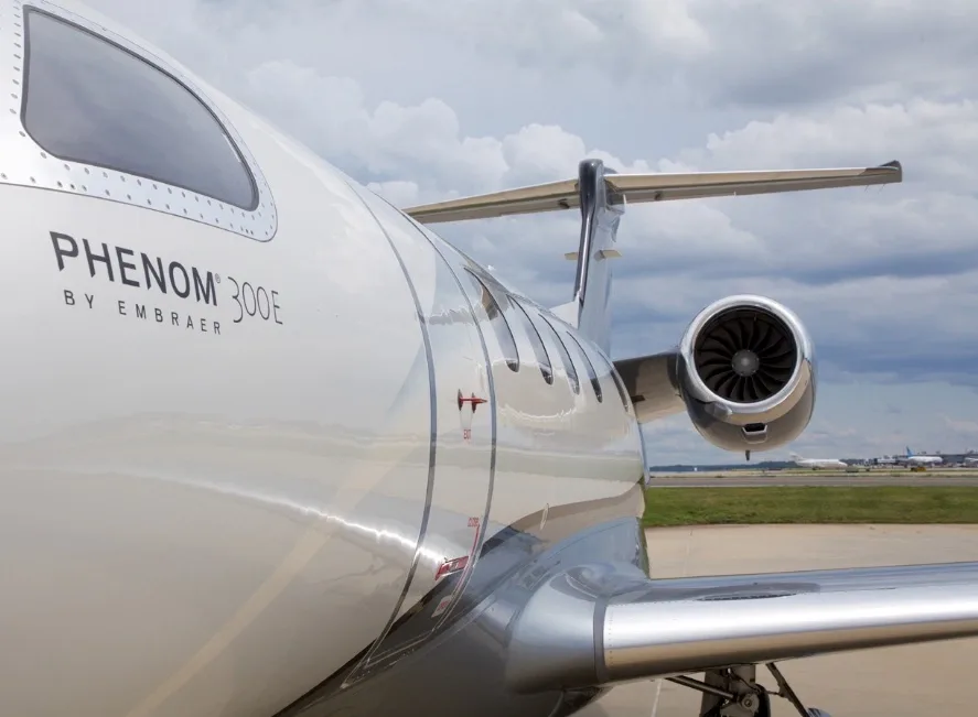 A close-up view of the fuselage and engine of an Embraer Phenom 300E, showing the "PHENOM 300E BY EMBRAER" text.