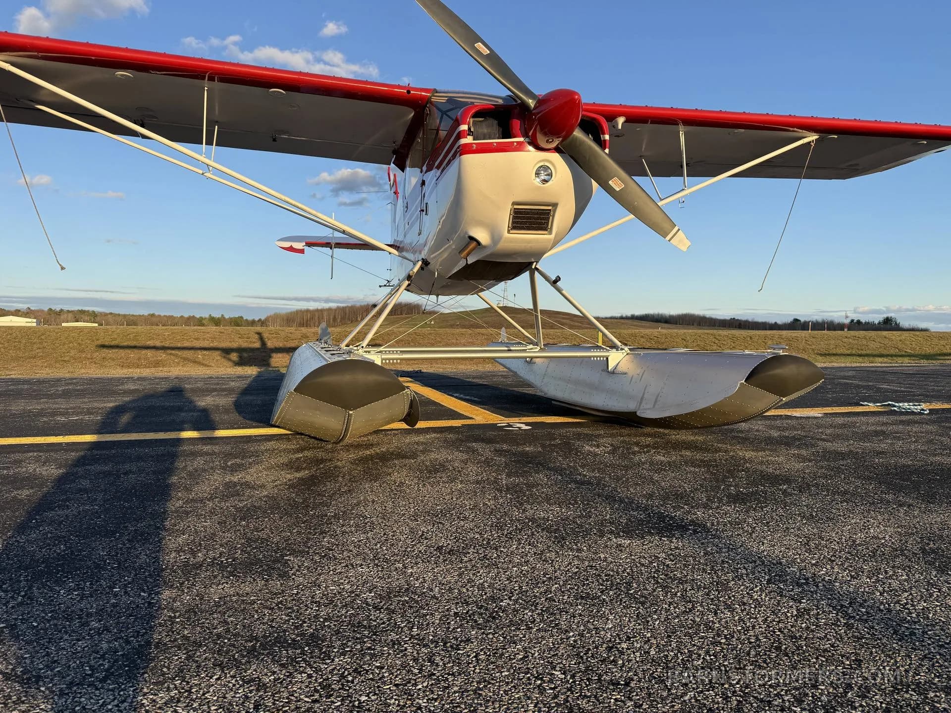 A low-angle front view of a red and white floatplane with a single propeller, parked on an asphalt runway.