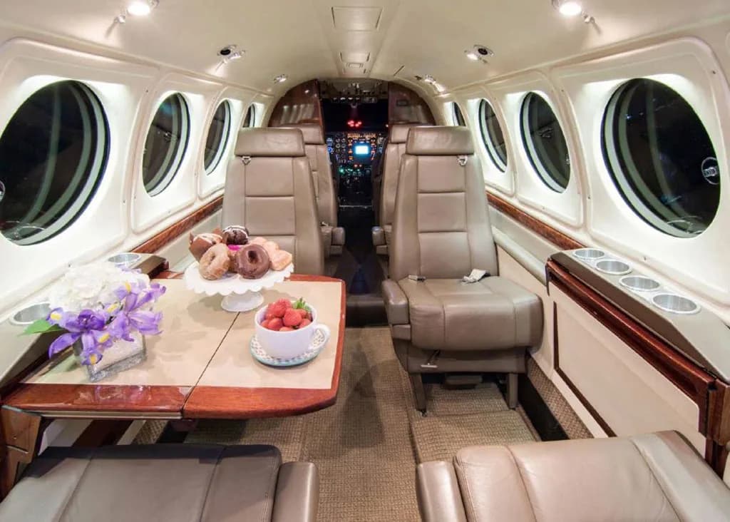The aircraft's interior cabin features beige leather seats, a wooden table with snacks and flowers, and round windows.