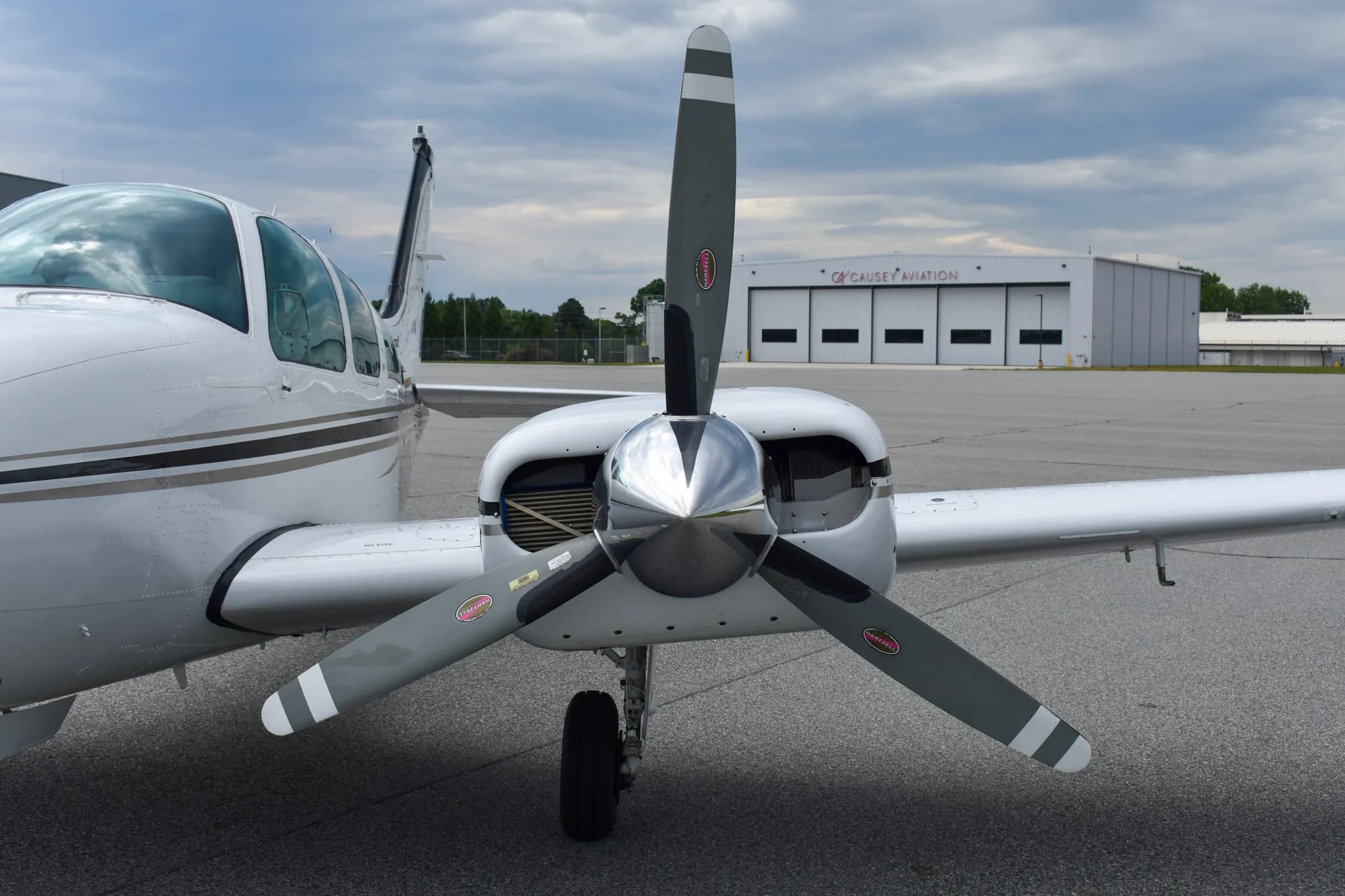 A front view of the left engine and propeller of a white Beechcraft Baron aircraft with hangars in the background.