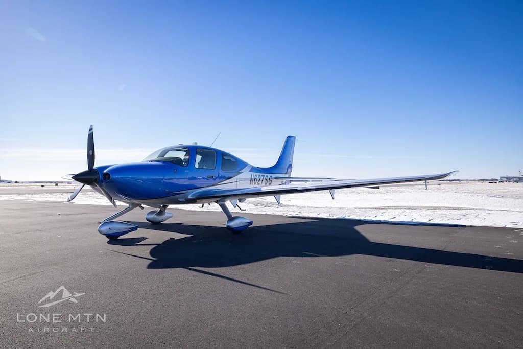 A blue and silver Cirrus SR22 aircraft (N627SG) parked on an airfield with snow on a sunny day.
