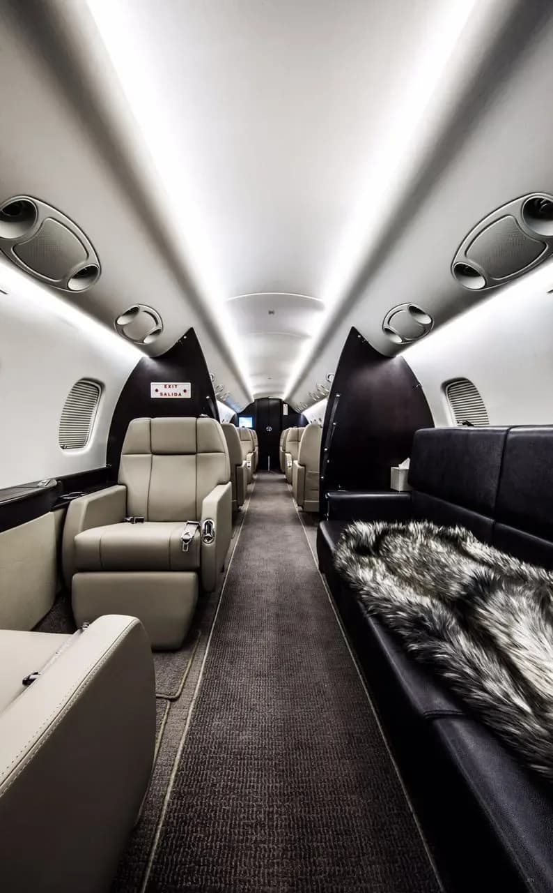 A long view of a luxurious private jet cabin aisle with beige seats and a dark couch with a fur throw.