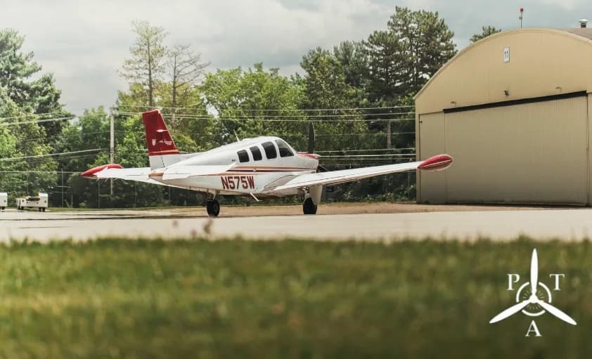 A white and red Beechcraft Bonanza aircraft, tail number N575W, seen from the side on a grassy airfield near a hangar.