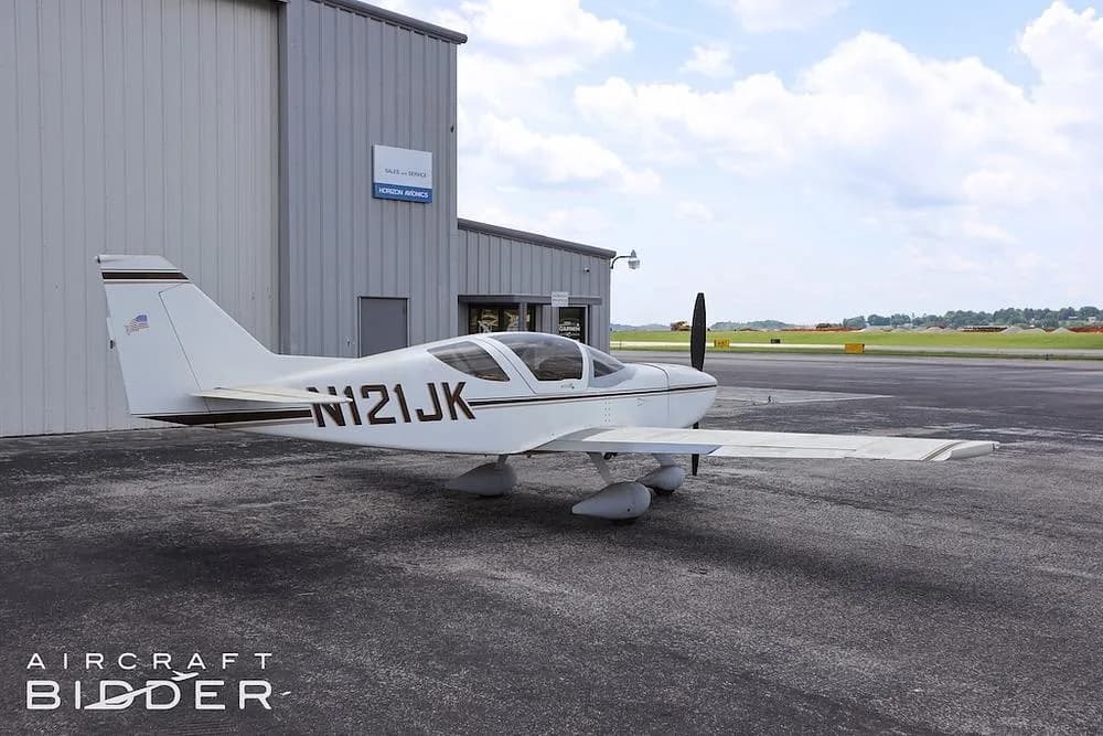 A white single-engine aircraft (N121JK) with brown stripes parked on an asphalt apron next to a hangar.