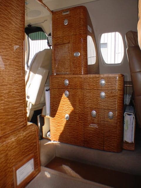 The interior cabin of a private jet, featuring light brown wood-grain cabinetry with silver handles and a view towards the front of the cabin.