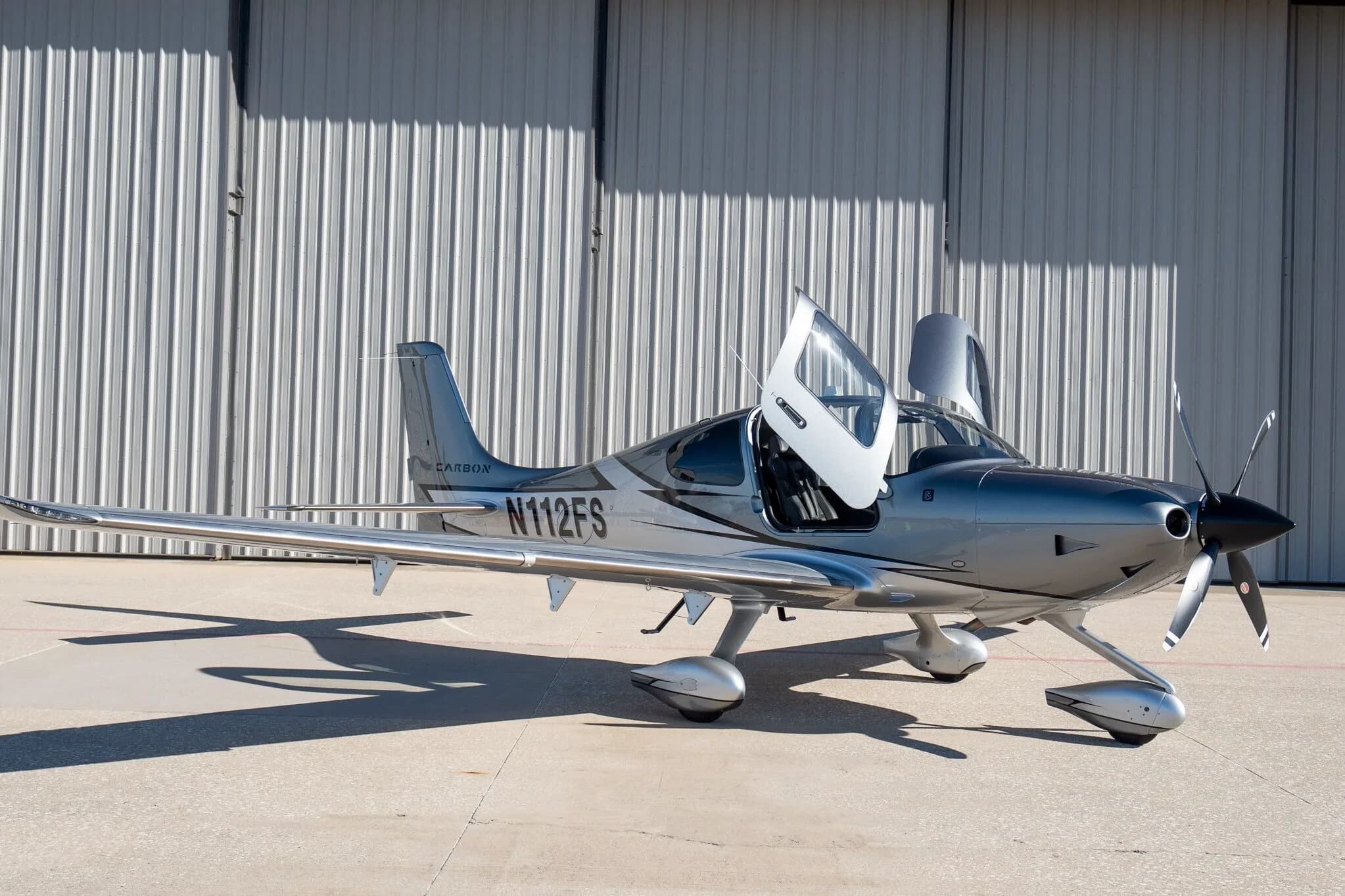 A front-right view of a silver and dark grey Cirrus SR22T Carbon Edition aircraft (N112FS) with its gull-wing doors open, parked in front of a hangar.