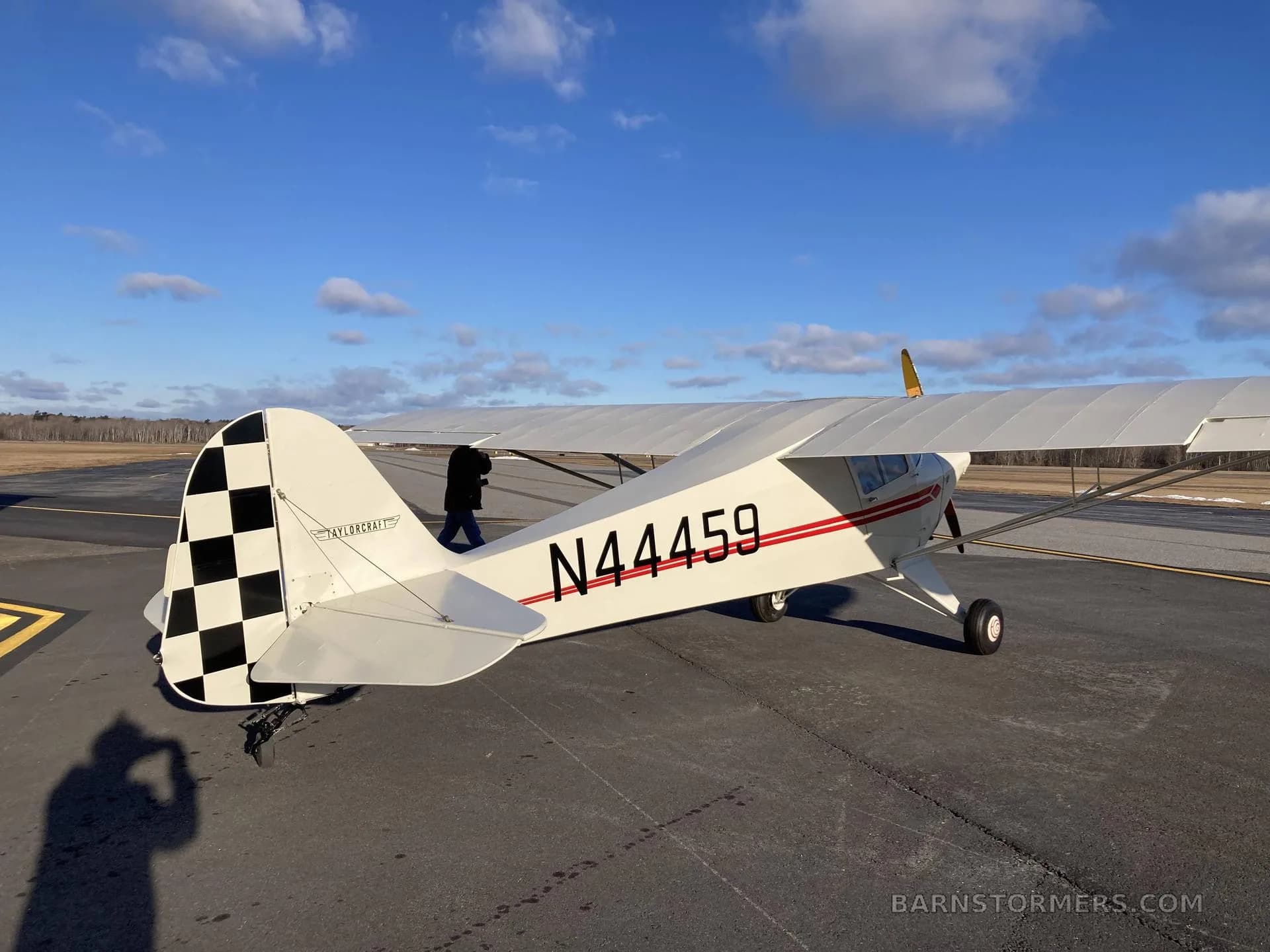 A rear quarter view of a white and red Piper Cub N44459 with a checkered tail, parked on an airfield.