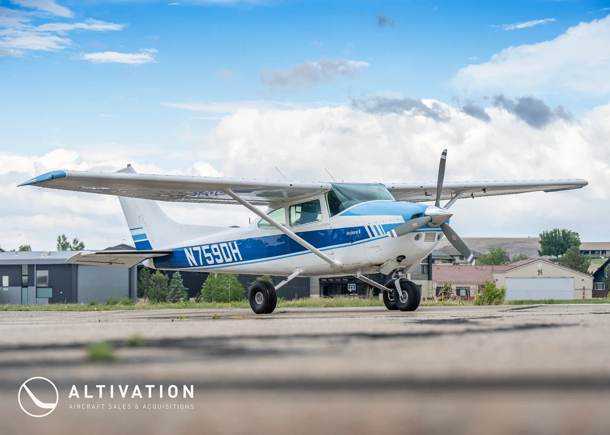 A white and blue Cessna 182 Skylane II (N759DH) parked on an airfield under a partly cloudy sky.