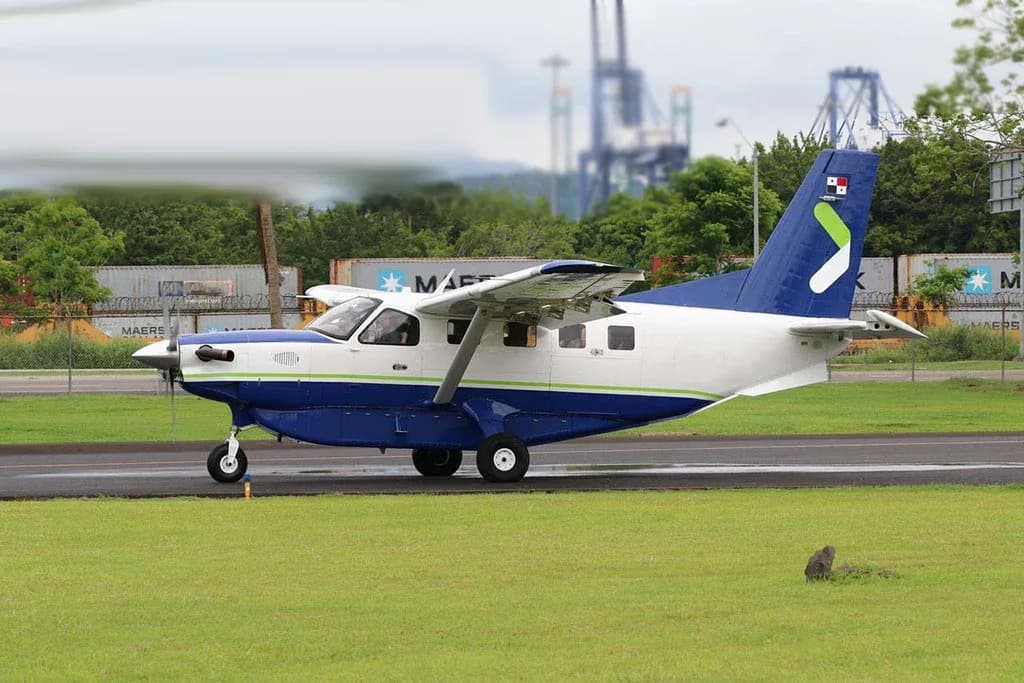 A blue and white Quest Kodiak 100 aircraft with a green stripe and a Panamanian flag on the tail, on a tarmac with industrial cranes in the background.