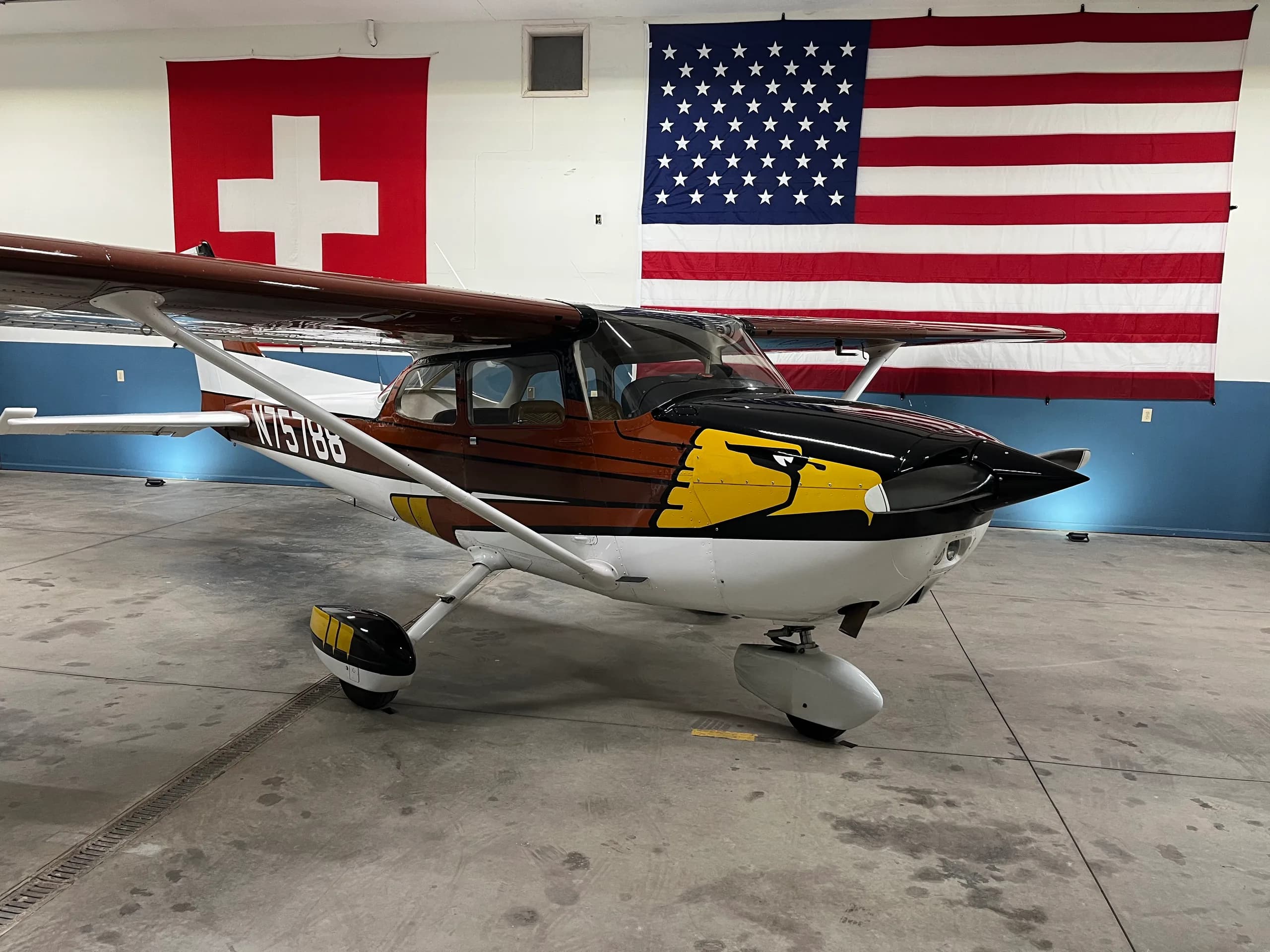 A brown and black Cessna-like aircraft with an eagle paint scheme on the nose, parked in a hangar with Swiss and American flags.