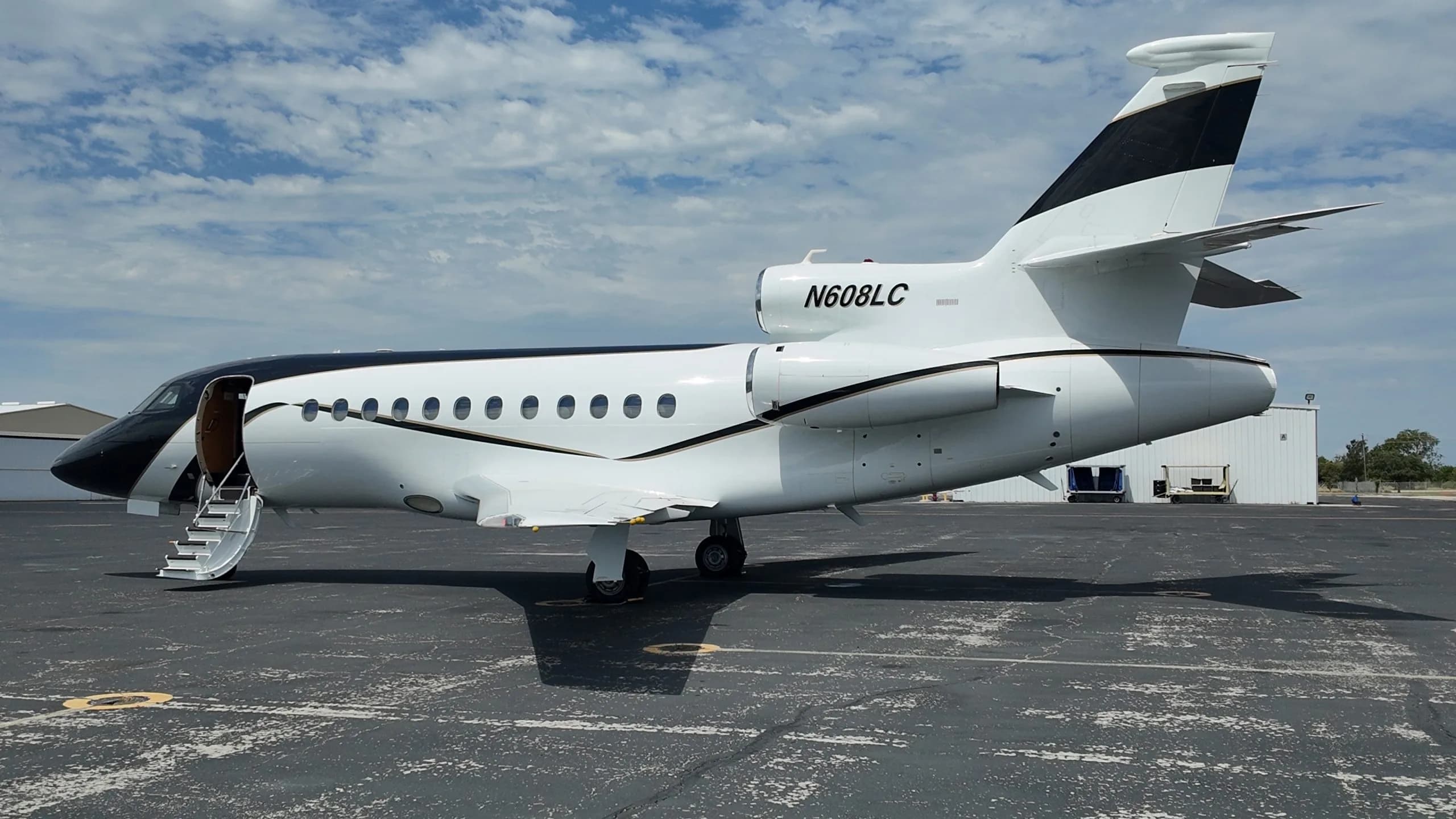 A side view of a white and black Dassault Falcon 50 business jet with its door open and stairs extended, parked on an asphalt tarmac under a partly cloudy sky.