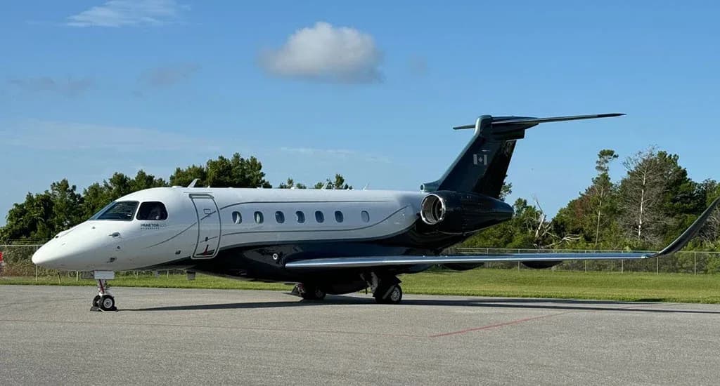 A white and dark grey Embraer Praetor 600 business jet with a Canadian flag on the tail, parked on an airfield under a blue sky.