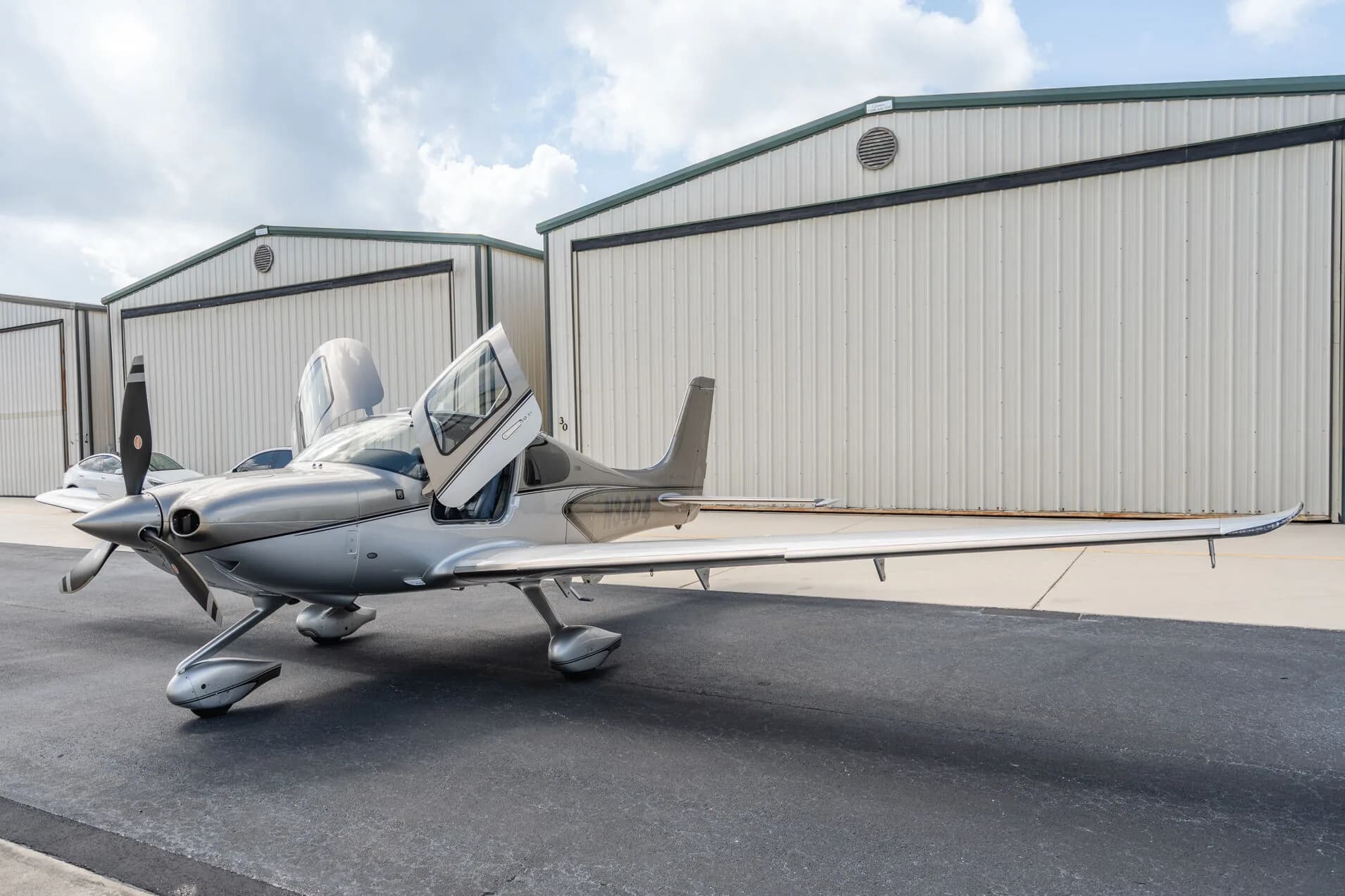Three-quarter front view of a silver Cirrus SR22T aircraft (N3404) with its gull-wing doors open, parked on an airport tarmac.