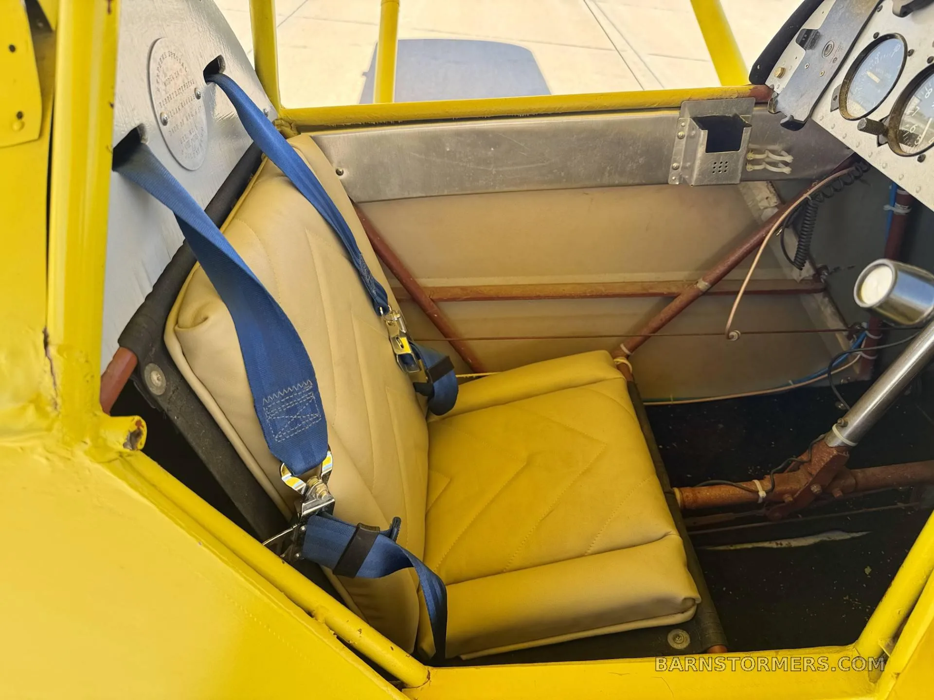 The single tan-colored seat with blue seatbelts inside the yellow biplane's open cockpit.