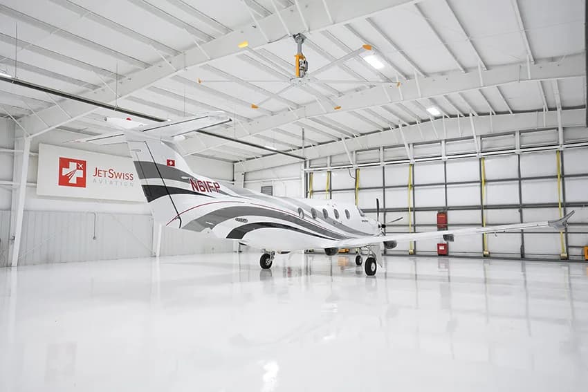A white Pilatus PC-12 aircraft with gray and red stripes is seen from the rear in a brightly lit hangar.