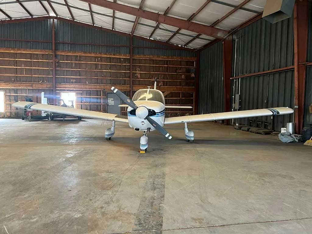 A head-on view of the white Piper Cherokee aircraft N53LA with blue stripes, parked inside a hangar.