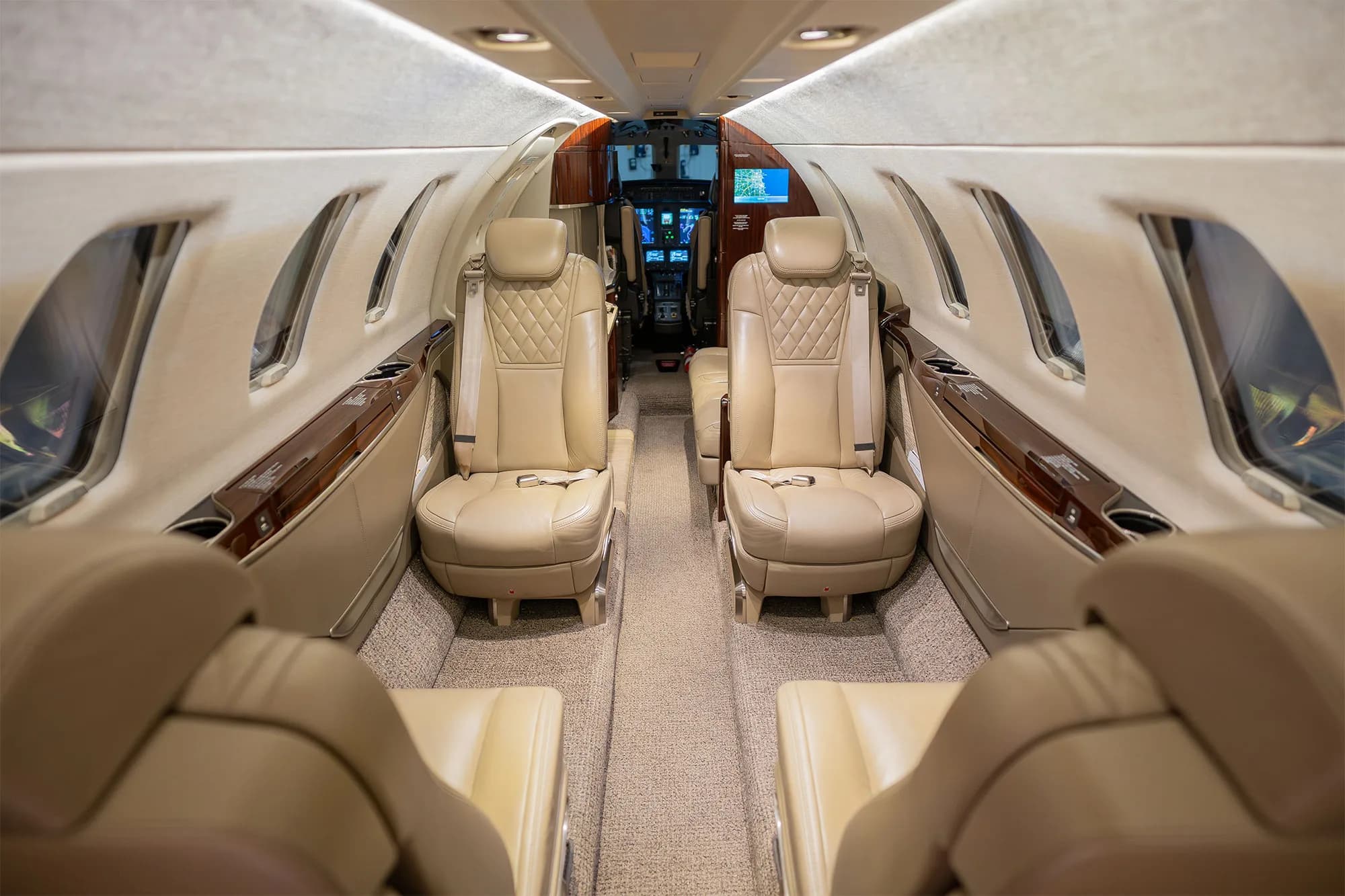 The luxurious interior cabin of a business jet, featuring beige leather seats and wood accents, looking towards the cockpit.