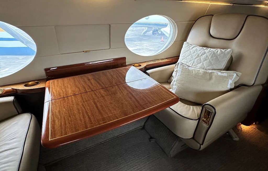 A close-up of a beige leather seat with pillows and a polished wooden table next to a window in a private jet cabin.