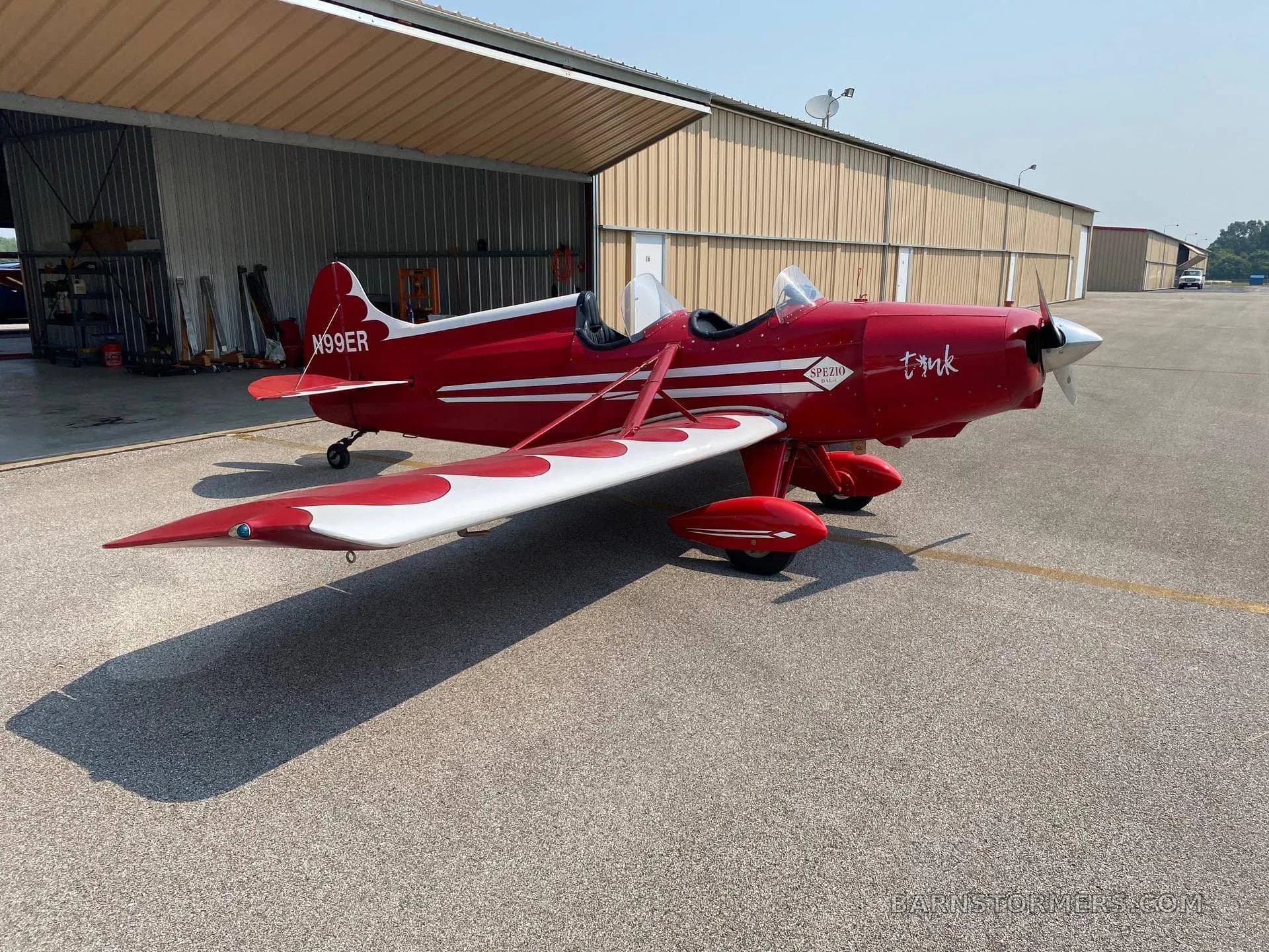 A red and white Spezio Tuholer monoplane, registration N99ER, parked on an asphalt apron in front of hangars on a sunny day.