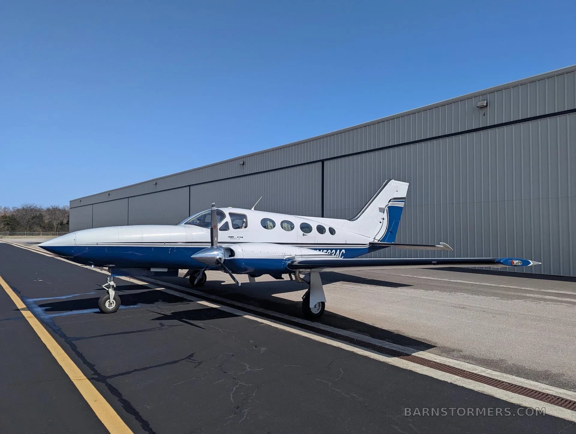 A white and blue twin-engine propeller aircraft (Cessna 414 Chancellor) with tail number N533AC parked on an asphalt ramp with yellow markings, viewed from the front-left side.