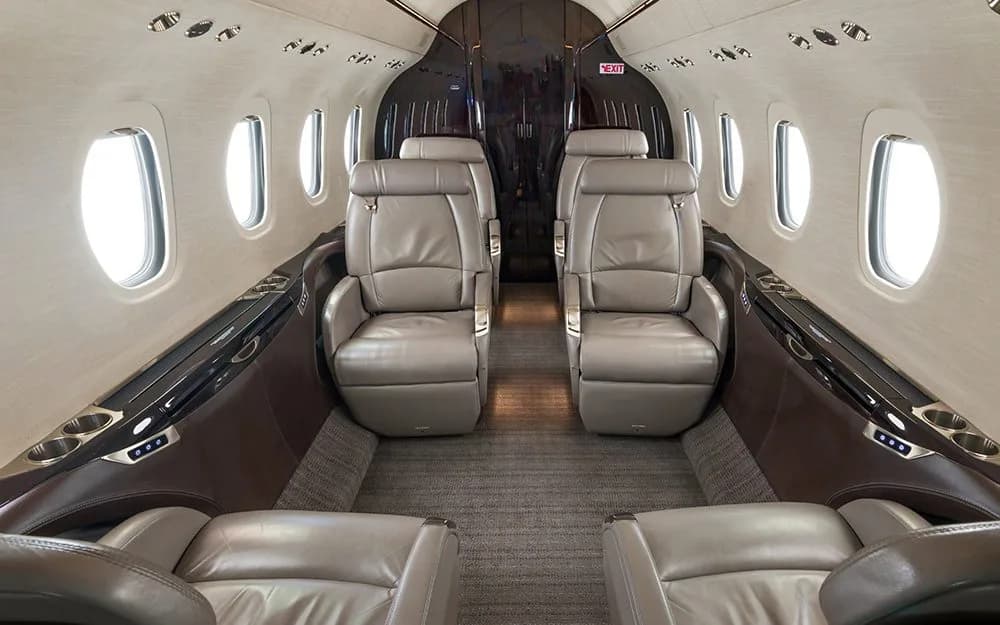 The interior cabin of a private jet, showing two rows of light brown leather seats facing each other.