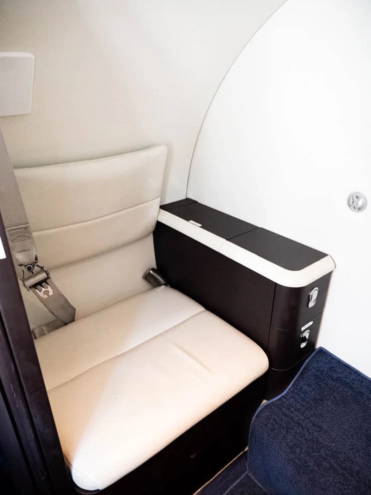 Aircraft interior with a white leather seat and dark wood storage unit.