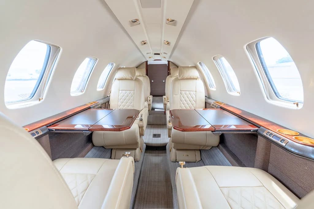 The interior cabin of a private jet showing light beige leather seats with deployed wooden fold-out tables.