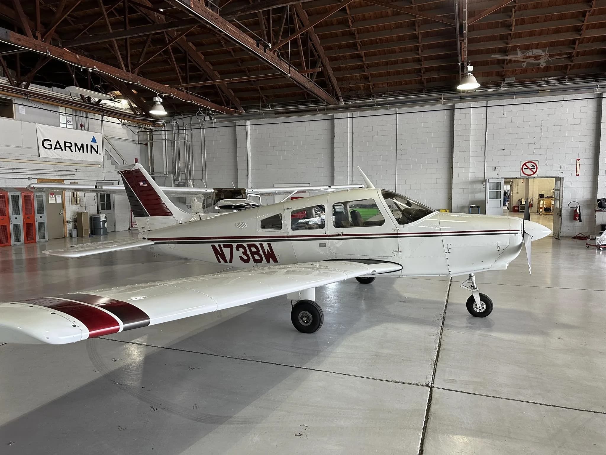 A white single-engine aircraft with maroon and black stripes, registration N73BW, viewed from the front-right in a hangar.