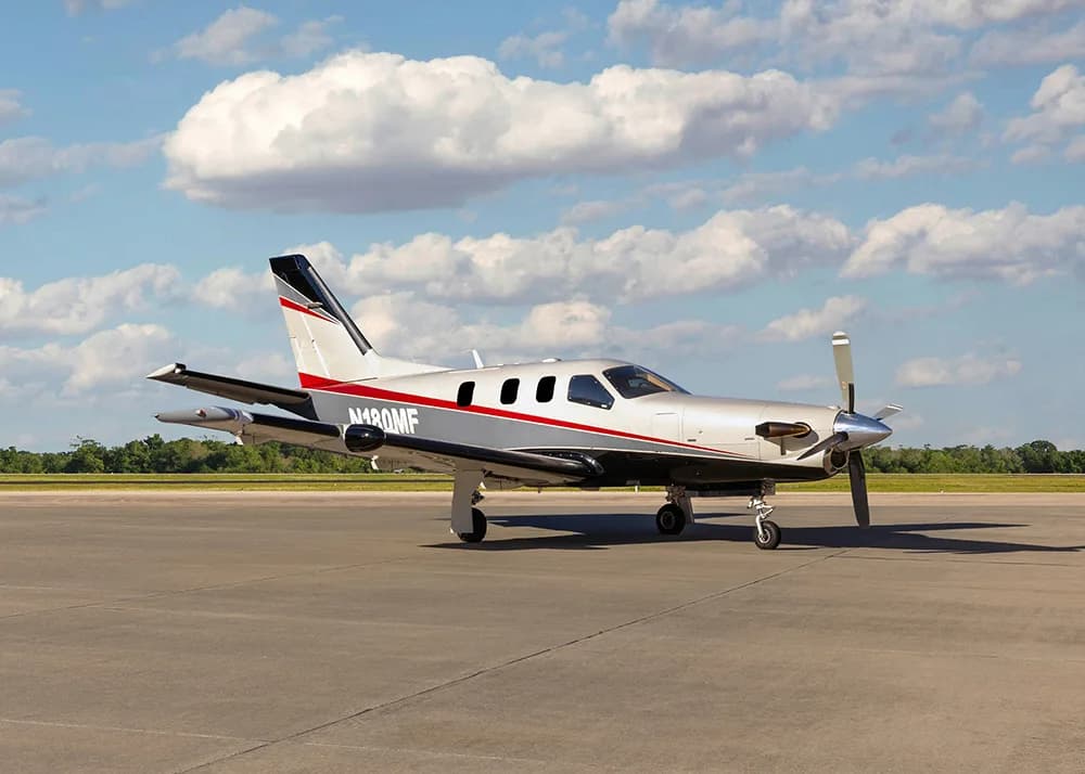 A white and grey TBM 900 series aircraft with red stripes, registration N180MF, parked on an airfield, viewed from the front-left with a slightly wider angle.